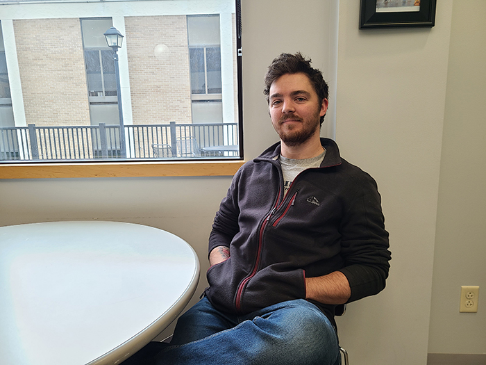 jon smith sitting at a table next to a gloomy looking window with his legs crossed and a smile on his face